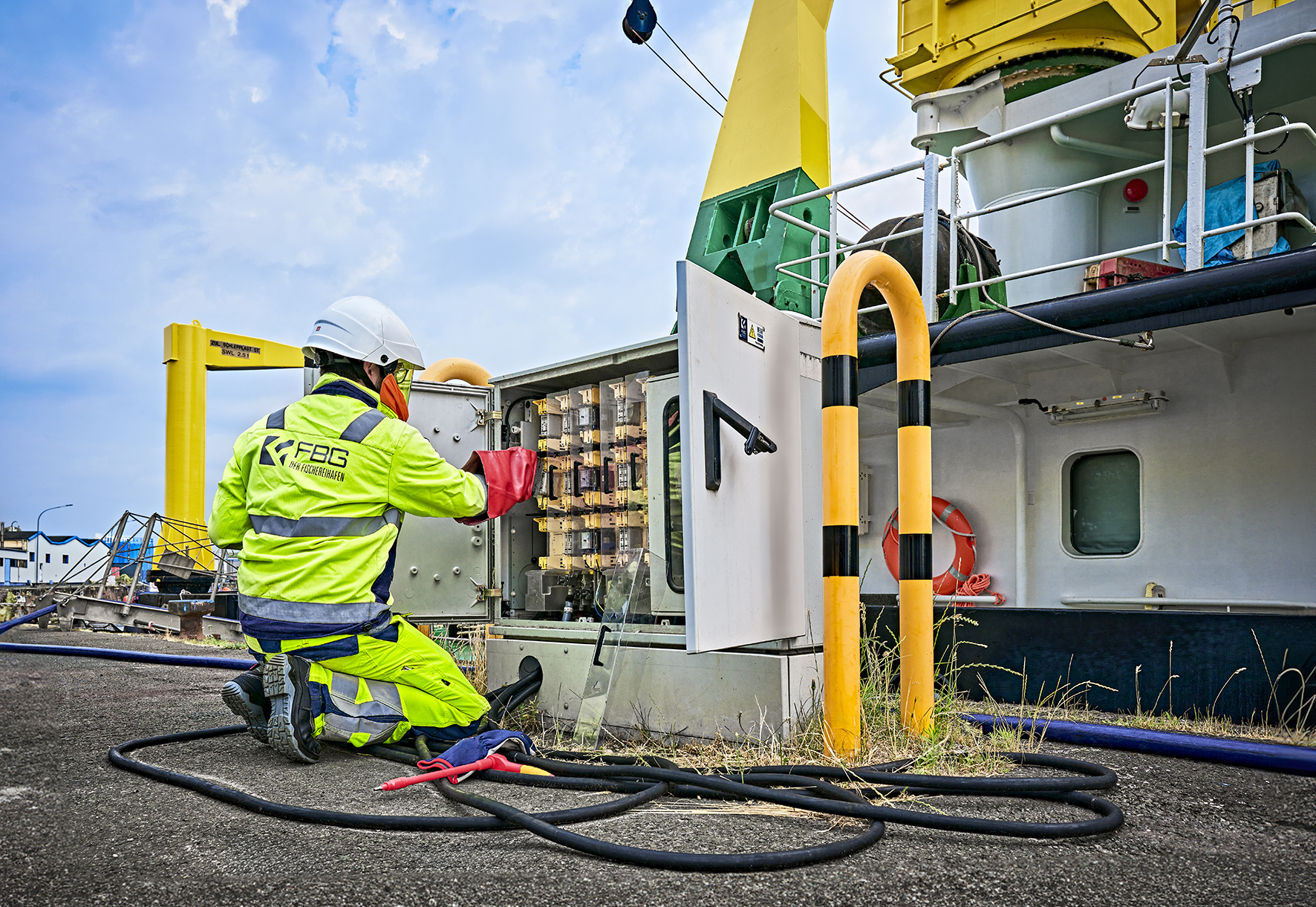 Das Bild zeigt einen Arbeiter in gelber Sicherheitskleidung und Helm, der an einem geöffneten Stromkasten arbeitet. Er kniet vor dem Kasten, der mit Kabeln und Schaltern verbunden ist, und trägt rote Schutzhandschuhe. Im Hintergrund befindet sich ein Schiff. Er kümmert sich um den Landstromanschluss für dieses Schiff.