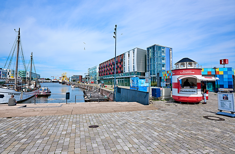 Im Schaufenster Fischereihafen Bremerhaven bei sonnigem Wetter. Links Schiff, rechts rot-weiß gestreifte Fischbrötchenbude, Tourist-Info und Hotel hinten. Von hier soll eine Hafenrundfahrt fahren.