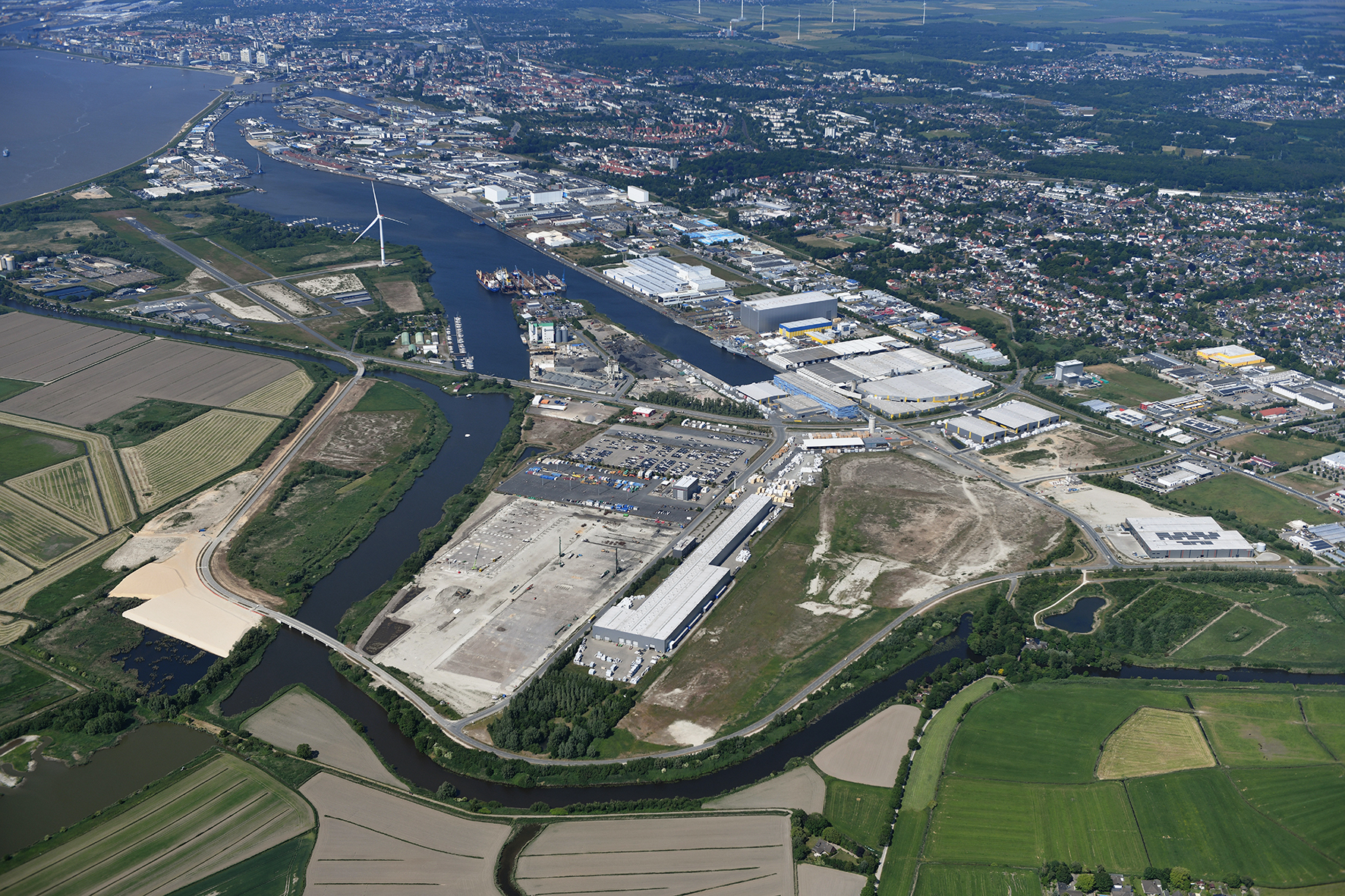 Das Bild zeigt eine Luftaufnahme des Fischereihafen Bremerhavens. der Fischereihafen ist ein weitläufiges Industrie- und Hafengebiet. Unten im Bild befinden sich grüne Felder und Wiesen. Darüber erstreckt sich das Hafengebiet mit großen Lagerhallen und Gebäudekomplexen. Windräder und einige Schiffe im Hafenbereich sind ebenfalls sichtbar. Am oberen rechten Rand erkennt man das Stadtgebiet von Bremerhaven.