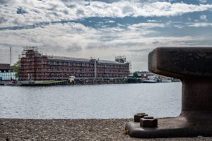 Backsteingebäude am Kai im Hintergrund, davor Wasser, Anlegepoller im Vordergrund, bewölkter Himmel.