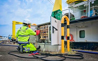 Das Bild zeigt einen Arbeiter in gelber Sicherheitskleidung und Helm, der an einem geöffneten Stromkasten arbeitet. Er kniet vor dem Kasten, der mit Kabeln und Schaltern verbunden ist, und trägt rote Schutzhandschuhe. Im Hintergrund befindet sich ein Schiff. Er kümmert sich um den Landstromanschluss für dieses Schiff.