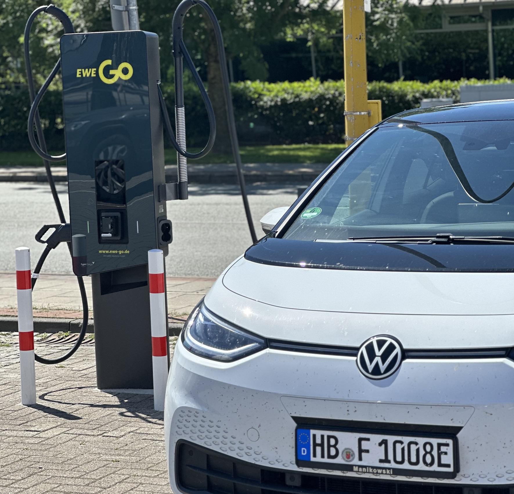 Das Bild zeigt ein weißes Elektroauto mit Bremer Kennzeichen, das an einer EWE Go Ladestation für Elektrofahrzeuge angeschlossen ist.