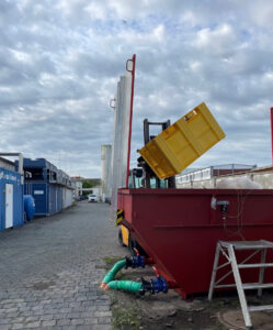 Ein Gabelstapler hebt eine gelbe Box und kippt sie in einen großen roten Container. Der Container ist mit grünen Schläuchen verbunden, die am Boden liegen. Im Hintergrund sind Industriegebäude und ein wolkiger Himmel zu sehen.