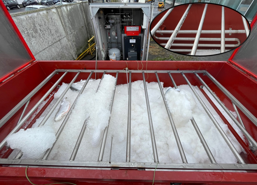 Roter Container mit zerkleinertem Eis, sogenannmtem Resteis, darüber Metallstangen, technische Anlage im Hintergrund. FBG und ttz untersuchen die Möglichkeiten zur Rückgewinnung von Kälteneergie aus dem Resteis.