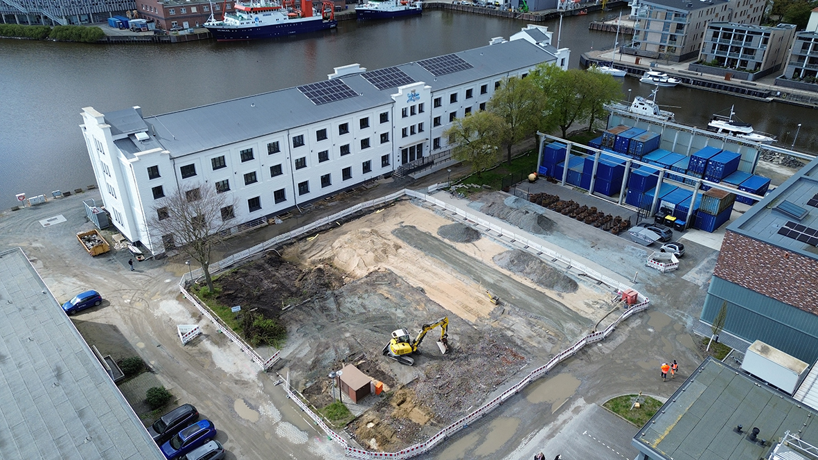 Baustelle vor einem großen, weißen Gebäude am Hafenbecken. Ein Bagger ist in einer ausgehobenen Fläche im Einsatz. Es handelt sich um die Freiflächen auf dem AWI-Campus, die nachhaltig gestaltet werden.