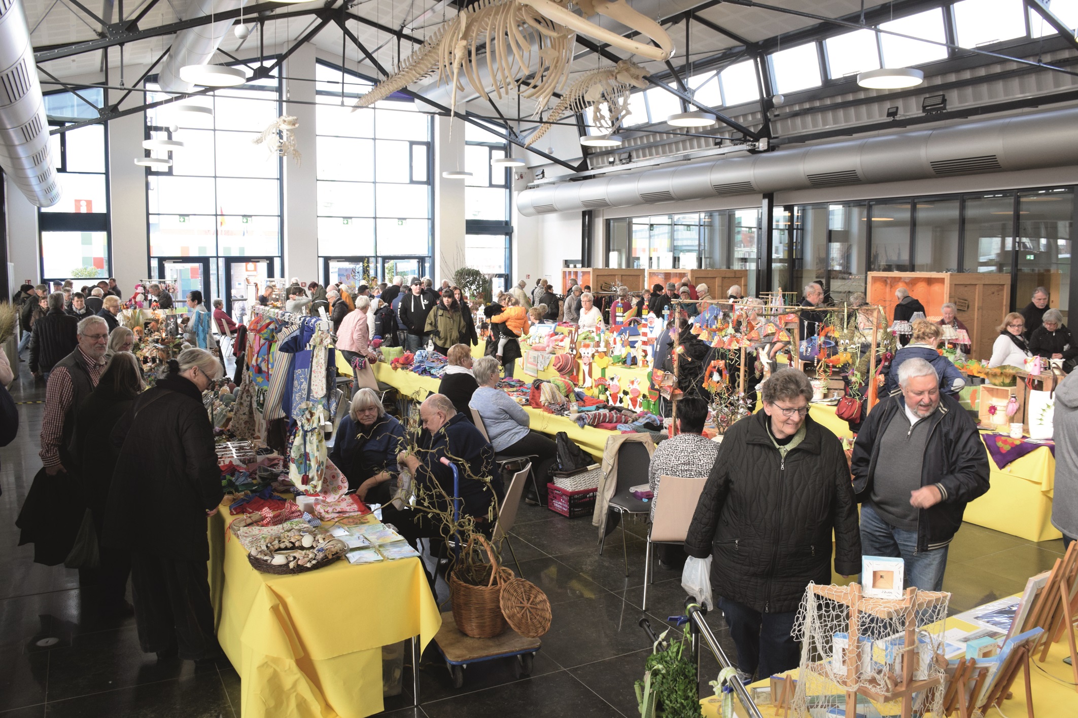 Das Bild zeigt einen belebten Kunsthandwerkermarkt in einer großen, lichtdurchfluteten Halle. Viele Menschen betrachten Stände mit bunten handgefertigten Waren. Über den Ständen hängt ein großes Skelett eines Meerestiers. Die Stimmung wirkt lebendig und geschäftig.