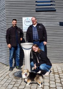 FBG Mitarbeitende und Ideengeber Waldemar Koch, Marco Staack, sowie Laura dos Santos und Hündin Bella posieren um den neuen Trinwasser-Spender.
